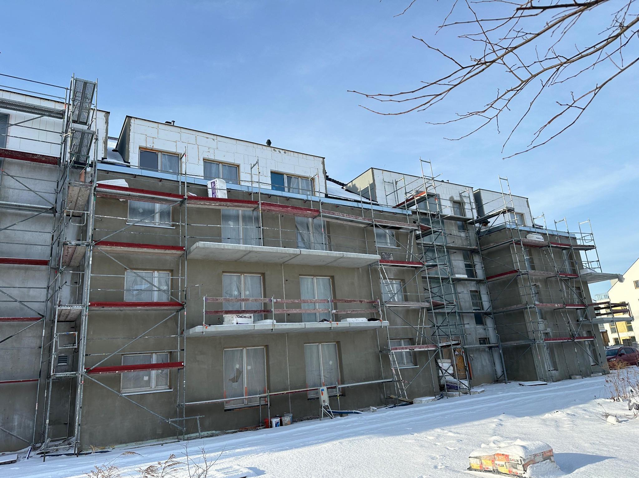 Wielopiętrowy budynek w trakcie prac elewacyjnych, otoczony rusztowaniami, częściowo ocieplony styropianem, widoczne worki z zaprawą na śniegu.