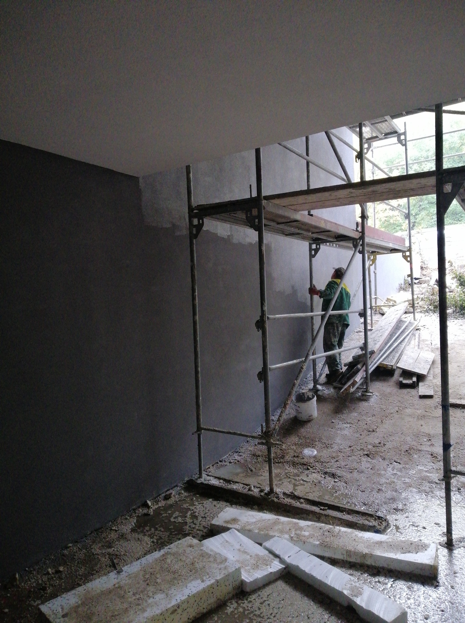 Pracownik w zielonym ubraniu na rusztowaniu, tynkuje szarą ścianę wewnątrz budynku, widoczne materiały budowlane i mokra podłoga.