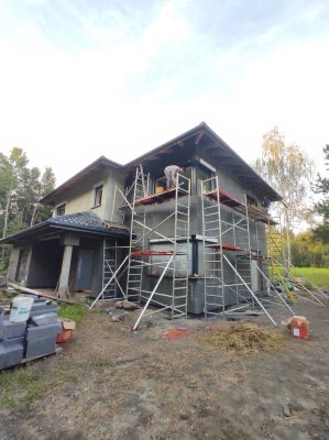 Dom w trakcie ocieplania elewacji, widoczne rusztowania, styropian i pracownik na wysokościach, przygotowanie do tynkowania.