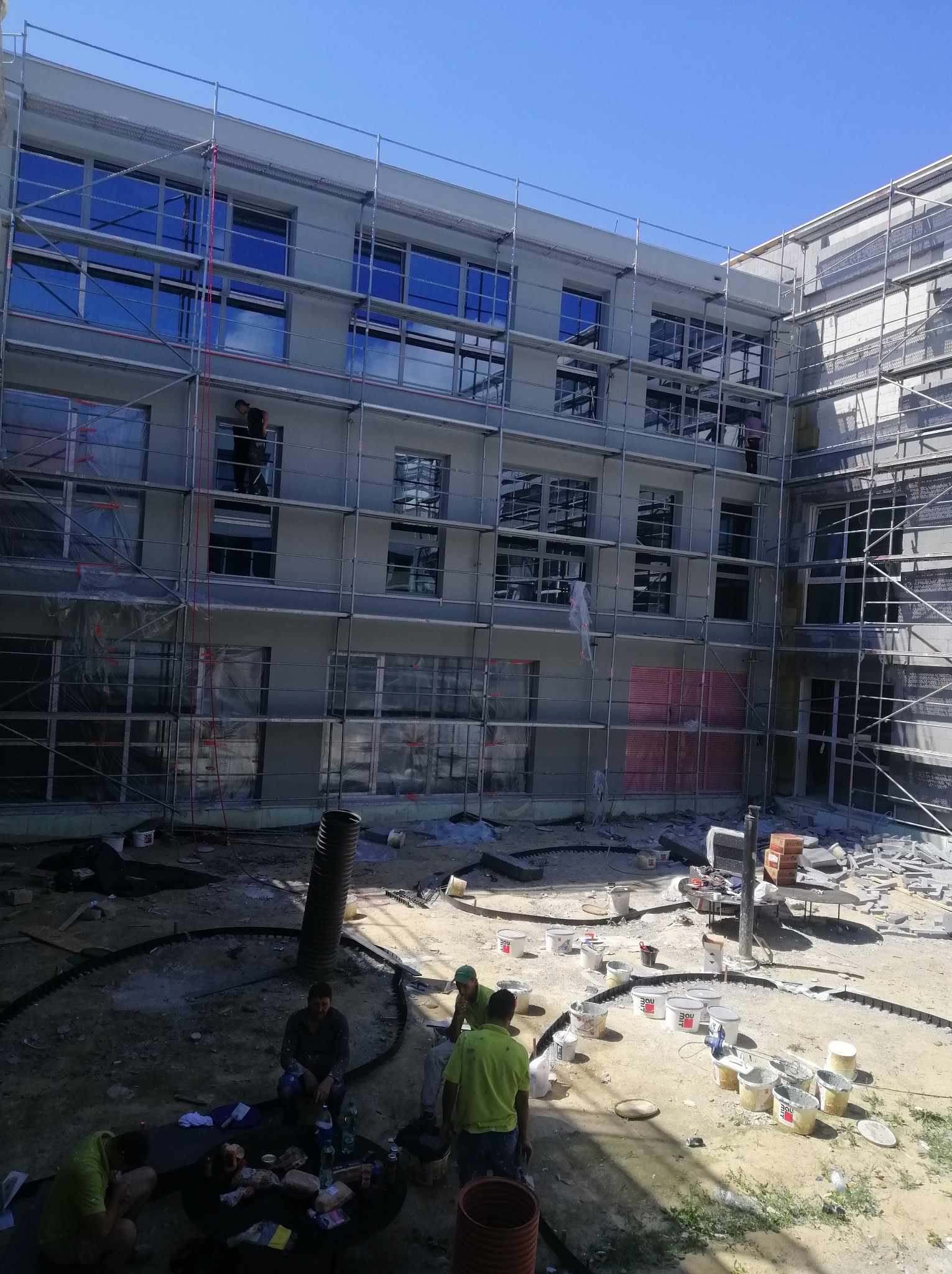 Budynek wielorodzinny w trakcie prac elewacyjnych, widoczne rusztowania, pracownicy na rusztowaniach oraz grupa pracowników odpoczywająca na ziemi, liczne wiadra i narzędzia budowlane w nieładzie.