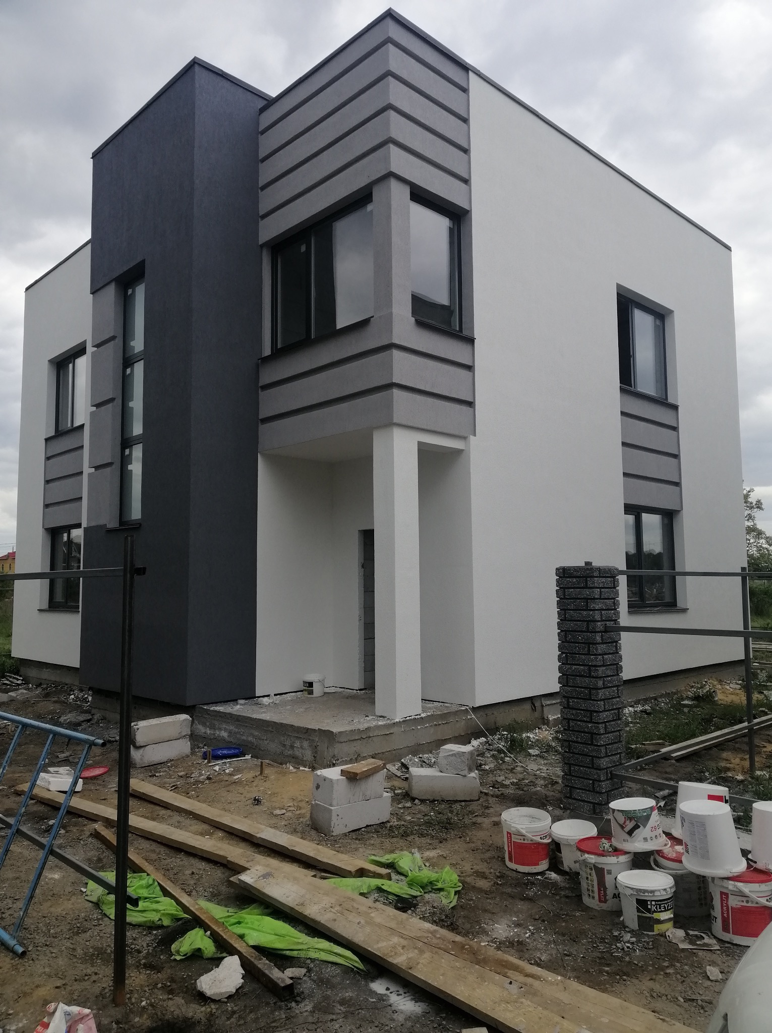 Nowoczesny, dwukondygnacyjny dom z elewacją w odcieniach szarości i bieli, widoczne puszki po farbie i materiały budowlane na posesji, pochmurne niebo w tle.