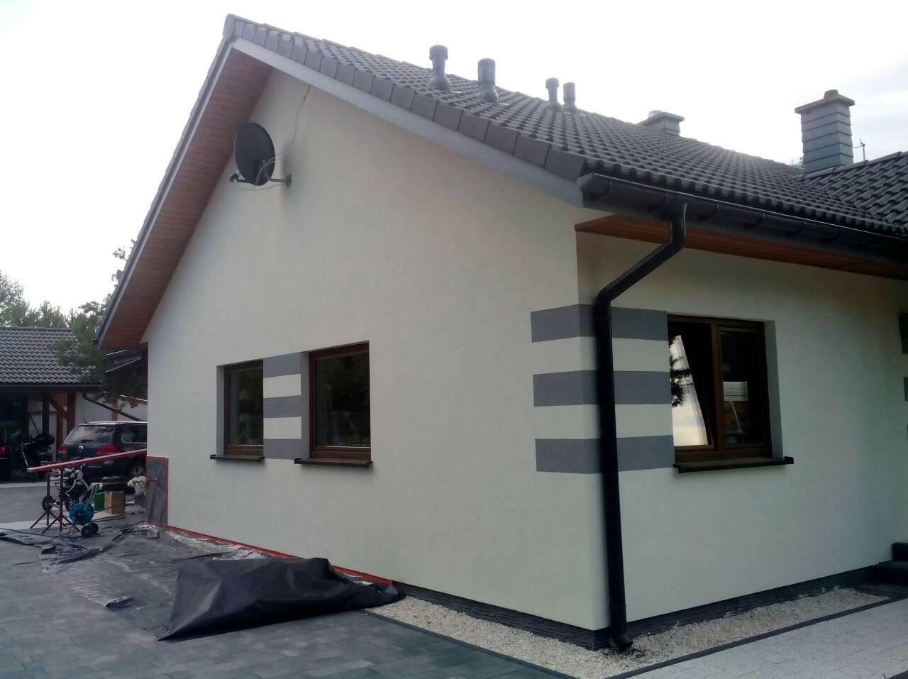 Ściana domu po tynkowaniu z poziomymi pasami szarej farby, widoczne okna z brązowymi ramami, dach z ciemnej dachówki, przygotowane narzędzia i folia ochronna na ziemi.