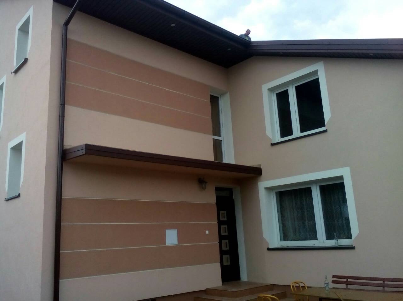 Wykończona elewacja domu z poziomymi pasami w odcieniach beżu i brązu, widoczne okna z białymi ramami i ciemny dach z rynną, na dachu widoczna sylwetka osoby.