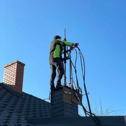 FREZ-STO Frezowanie i renowacja kominów - Mężczyzna w zielonej kurtce, stojący na rusztowaniu zamontowanym na kominie, wykonuje prace na wysokości z użyciem specjalistycznego sprzętu.