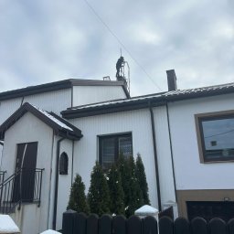FREZ-STO Frezowanie i renowacja kominów - Osoba w ciemnym kombinezonie na drabinie dachowej, prawdopodobnie czyszcząca komin na tle pochmurnego nieba i białego domu z brązowymi akcentami.