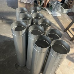 FREZ-STO Frezowanie i renowacja kominów - Grupa pionowo ustawionych, nowych, metalowych rur wentylacyjnych o cylindrycznym kształcie, z widocznymi oznaczeniami pisakiem, w surowym pomieszczeniu.