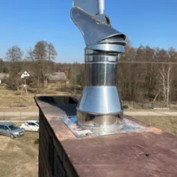 Błyszcząca, metalowa nasada kominowa na dachu z cegły, widok z góry na okolicę z domami i drzewami w słoneczny dzień.