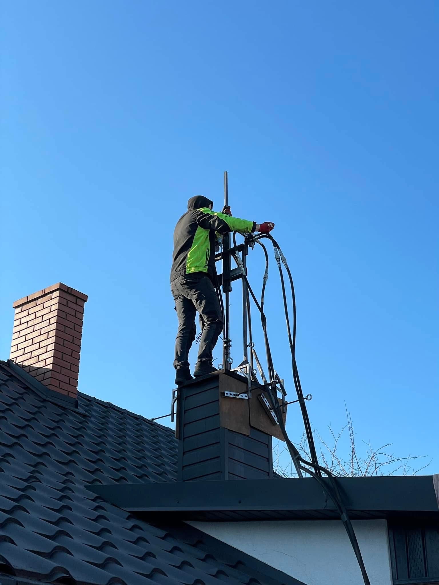 Mężczyzna w zielonej kurtce, stojący na rusztowaniu zamontowanym na kominie, wykonuje prace na wysokości z użyciem specjalistycznego sprzętu.