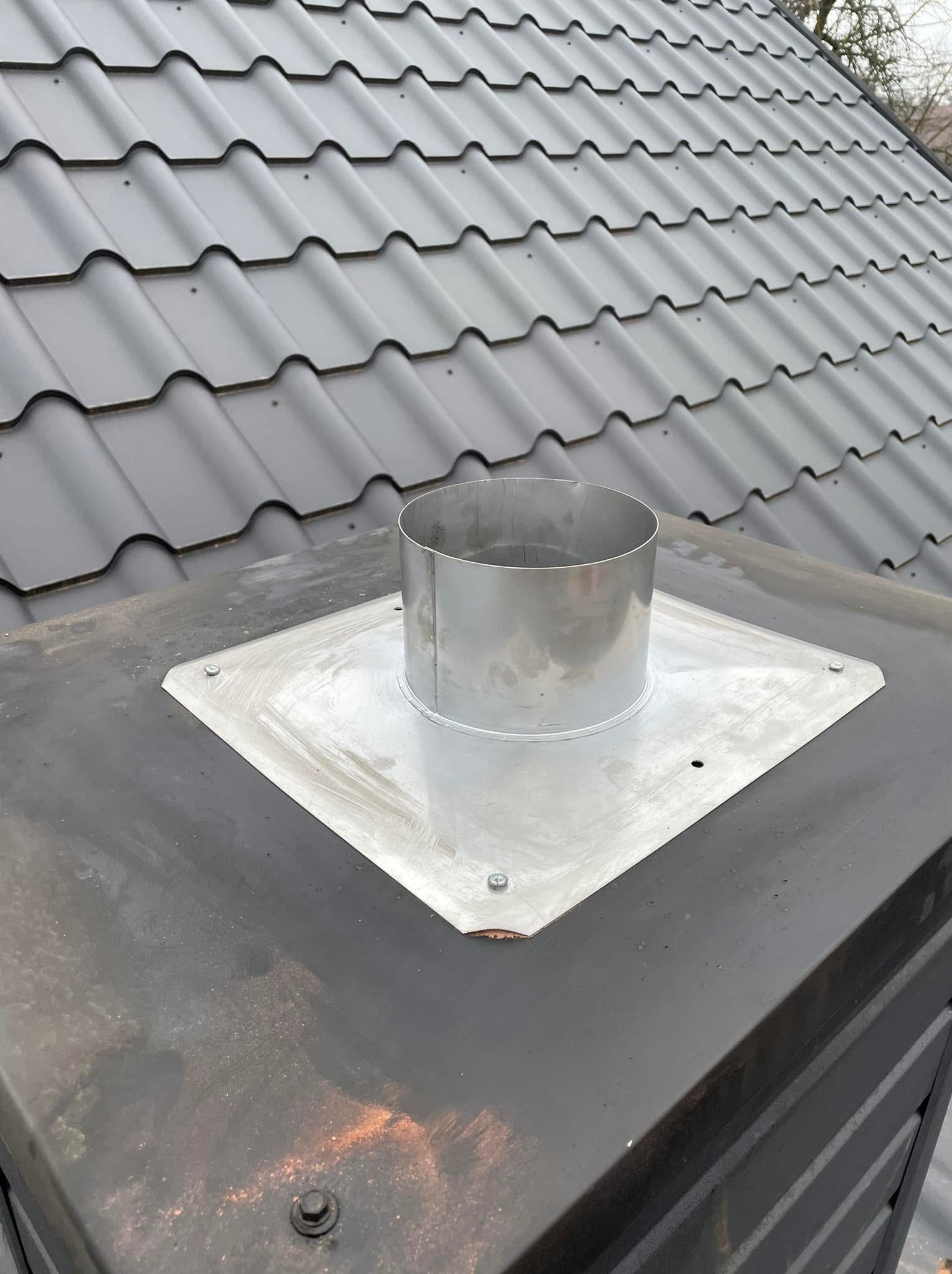 Metalowy komin z kołnierzem uszczelniającym na dachu z blachodachówki w kolorze grafitowym, widoczne ślady spawania i mocowania.