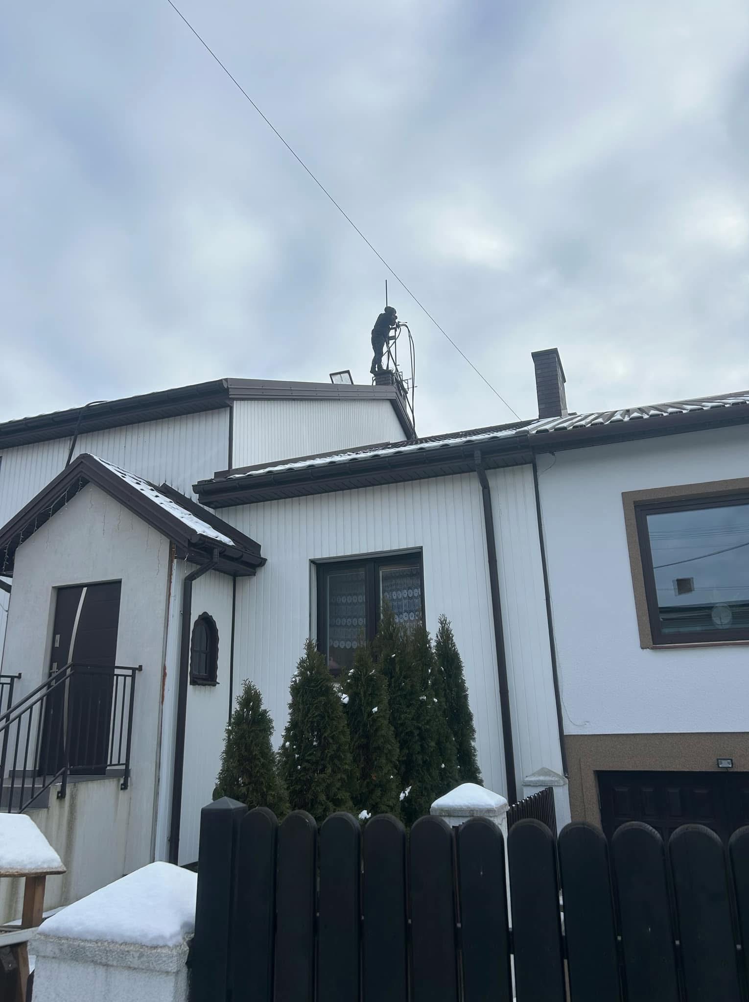 Osoba w ciemnym kombinezonie na drabinie dachowej, prawdopodobnie czyszcząca komin na tle pochmurnego nieba i białego domu z brązowymi akcentami.
