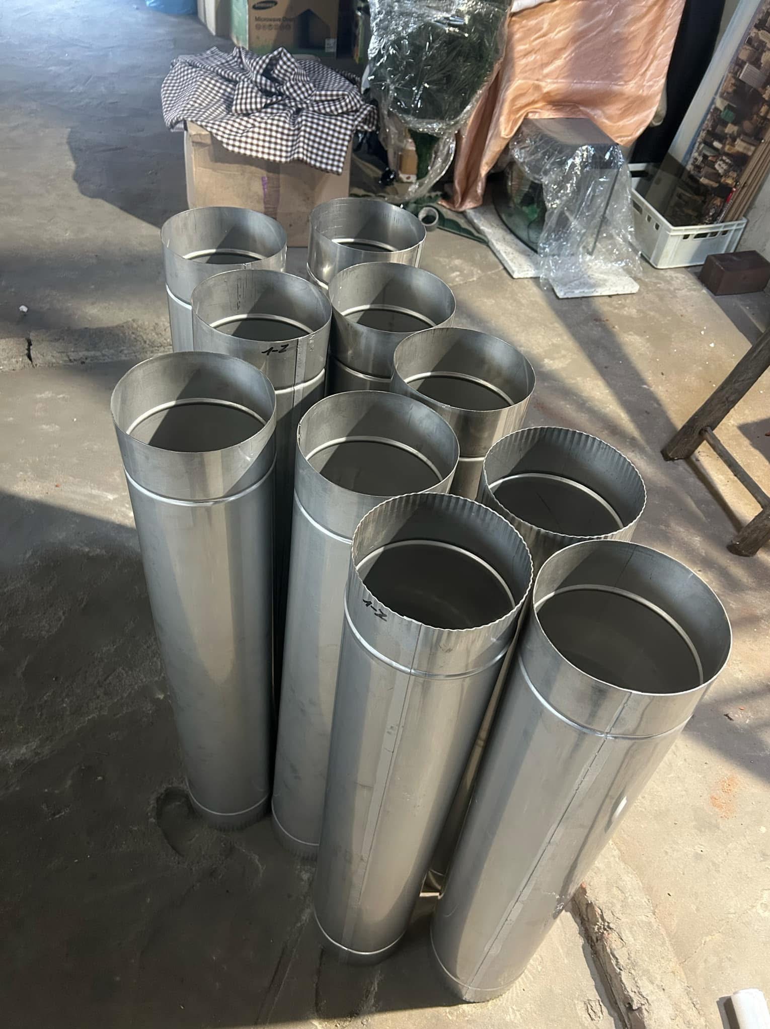 Grupa pionowo ustawionych, nowych, metalowych rur wentylacyjnych o cylindrycznym kształcie, z widocznymi oznaczeniami pisakiem, w surowym pomieszczeniu.