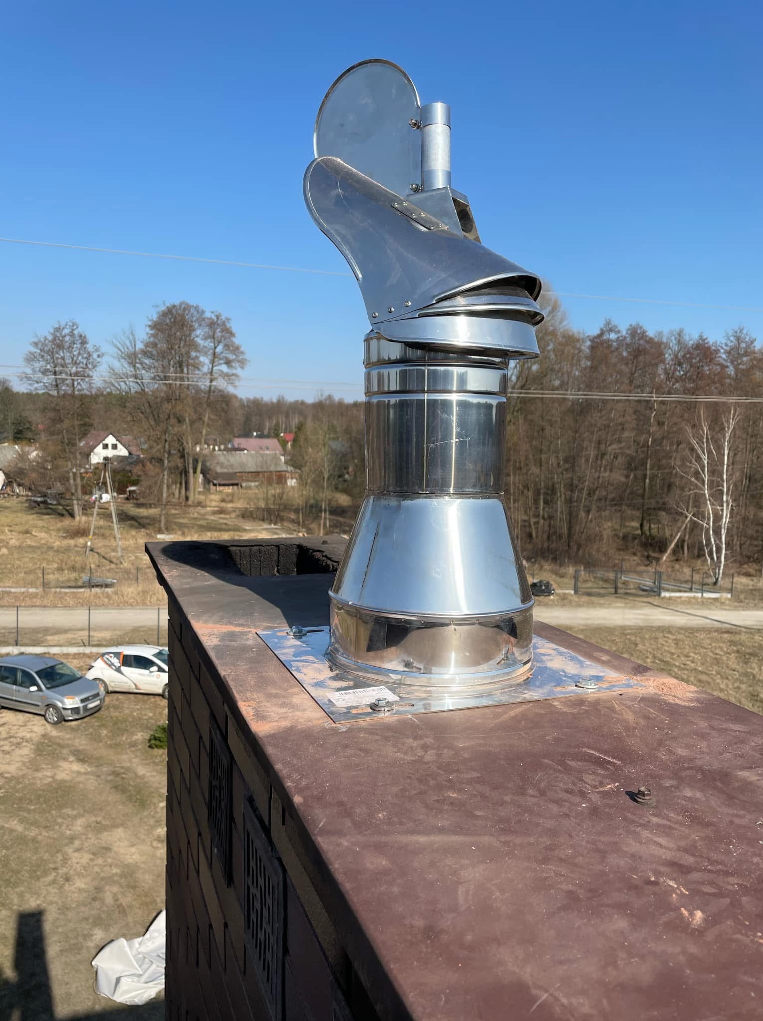 Błyszcząca, metalowa nasada kominowa na dachu z cegły, widok z góry na okolicę z domami i drzewami w słoneczny dzień.