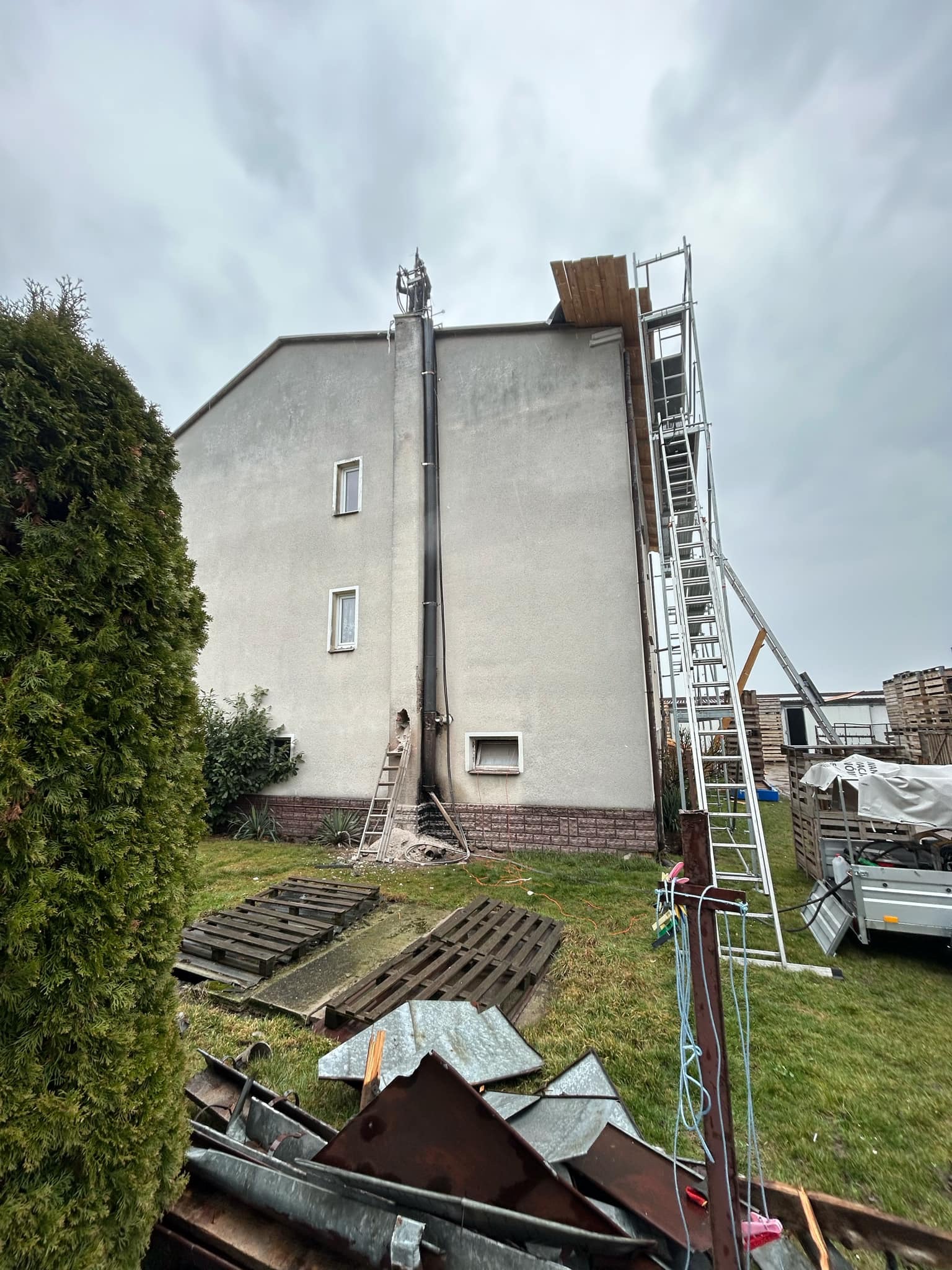 Montaż rury spalinowej na elewacji budynku jednorodzinnego, widoczny rusztowanie i drabina, pozostałości po kuciu w ścianie oraz składowane materiały budowlane na trawniku.