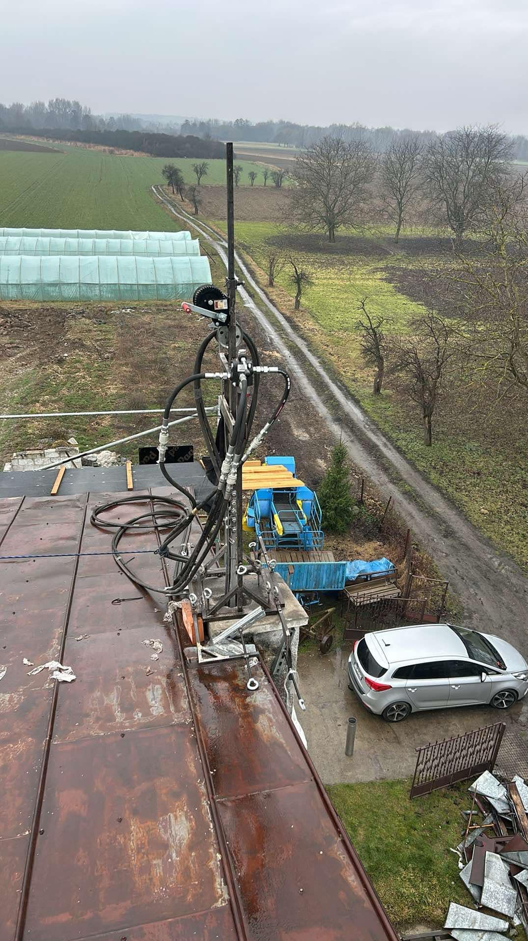 Widok z dachu na wieś, z zamontowanym urządzeniem do prac wysokościowych z czarnymi wężami, na tle pól, szklarni i drogi gruntowej, pochmurny dzień.
