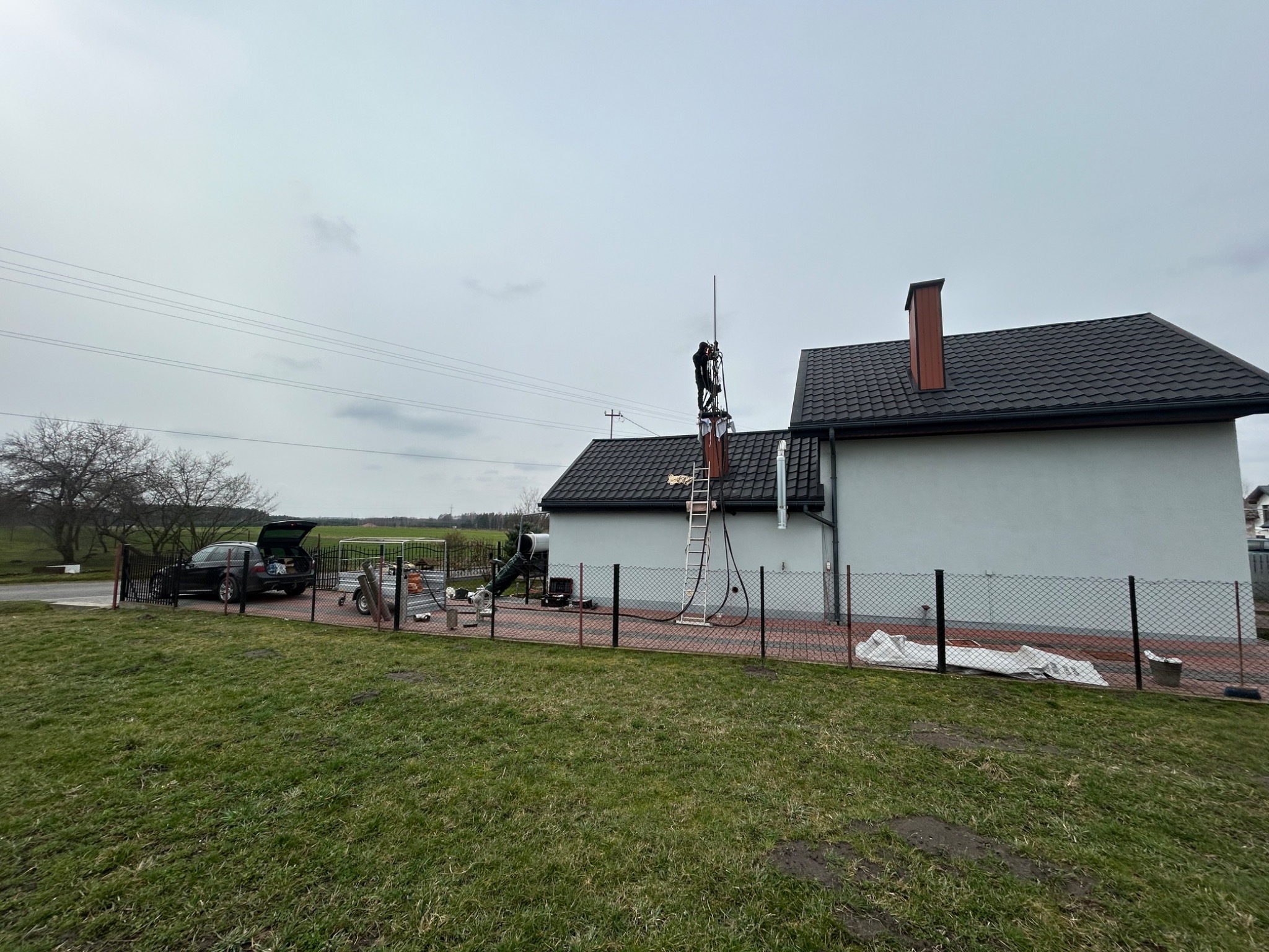 Pracownik na drabinie przy kominie domu jednorodzinnego, montaż lub serwis instalacji kominowej, widoczny samochód z przyczepką i narzędzia na posesji.