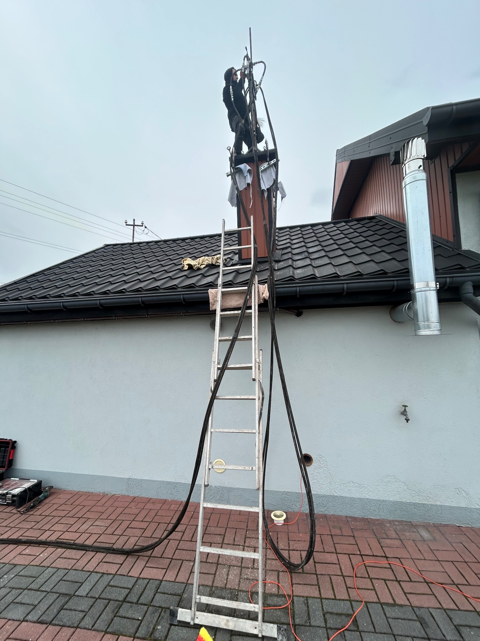 Mężczyzna w czarnym kombinezonie na szczycie drabiny, czyszczący komin z użyciem specjalistycznego sprzętu; widoczne narzędzia i przewody na dachu oraz przy ścianie budynku.