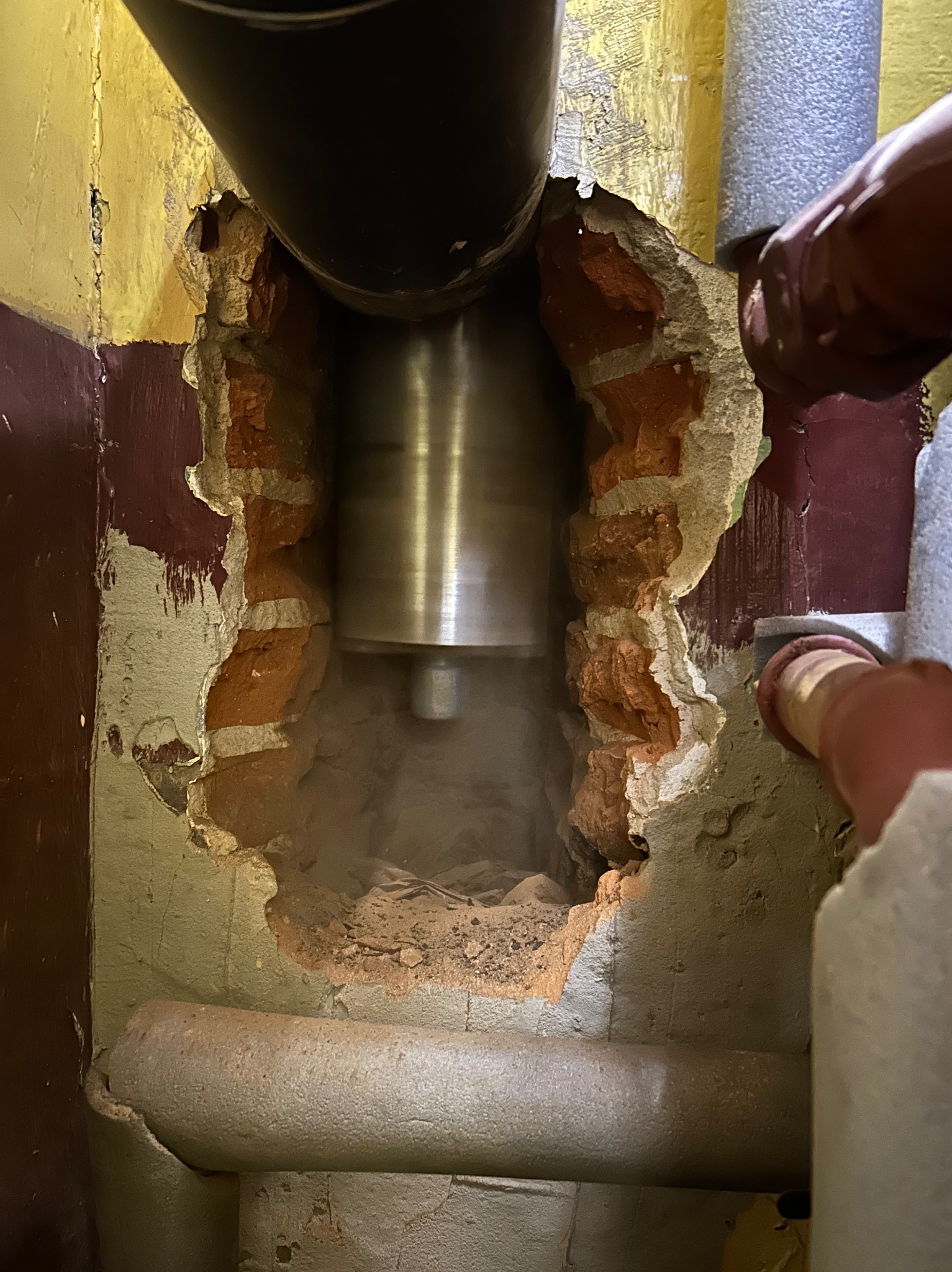 Wiercenie rdzeniowe w ceglanej ścianie podczas montażu rury wentylacyjnej, widoczne ślady pyłu i uszkodzenia tynku.