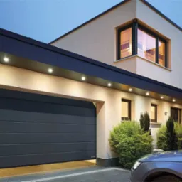 Nowoczesna, segmentowa brama garażowa w kolorze antracytowym, częściowo otwarta, z oświetleniem LED w podsufitce, widoczna fasada domu z oknami i numerem 67, fragment samochodu na pierwszym planie.