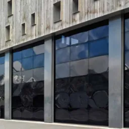 Nowoczesna, czarna brama przemysłowa segmentowa z metalowymi prowadnicami, zamontowana w budynku o drewnianej elewacji z licznymi, małymi oknami.