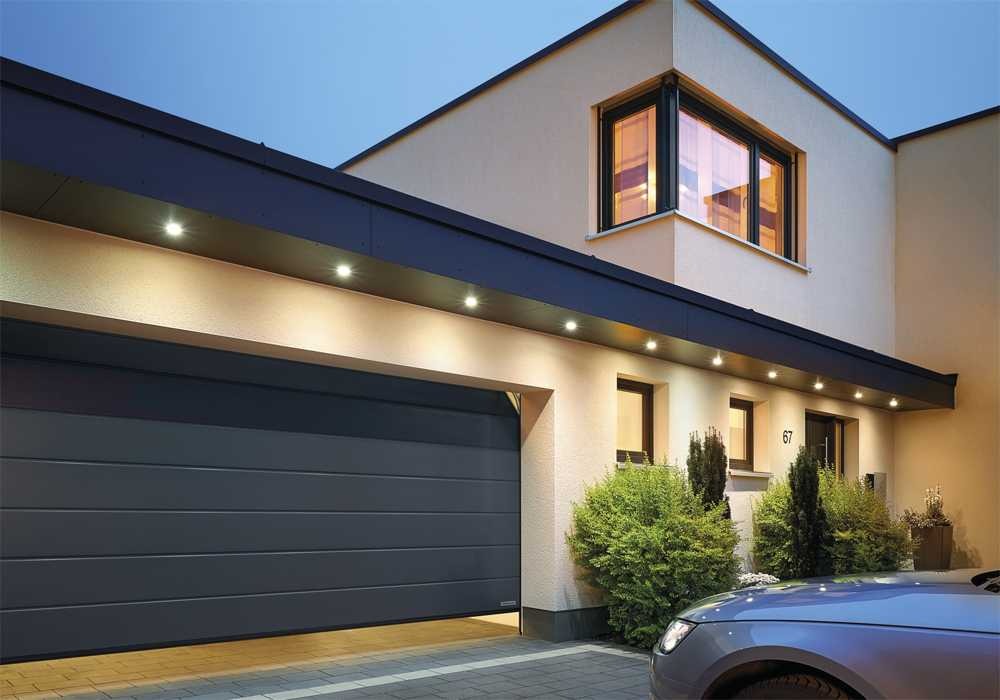 Nowoczesna, segmentowa brama garażowa w kolorze antracytowym, częściowo otwarta, z oświetleniem LED w podsufitce, widoczna fasada domu z oknami i numerem 67, fragment samochodu na pierwszym planie.