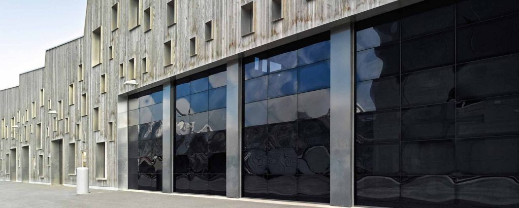 Nowoczesna, czarna brama przemysłowa segmentowa z metalowymi prowadnicami, zamontowana w budynku o drewnianej elewacji z licznymi, małymi oknami.