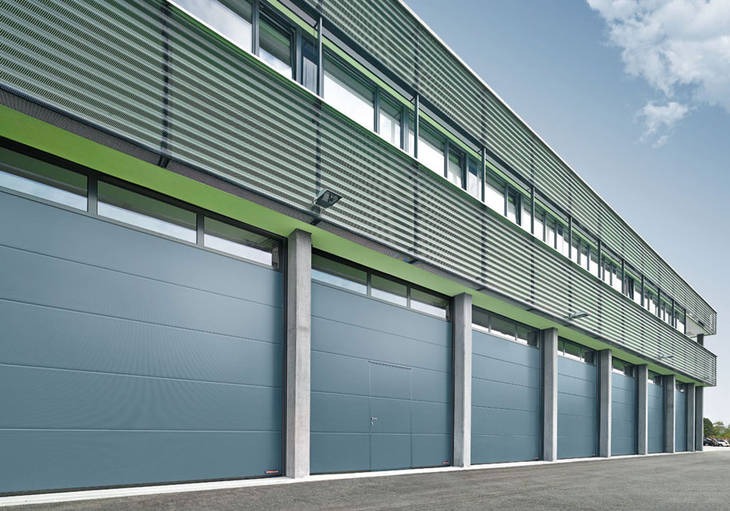 Szereg szarych bram garażowych segmentowych z oknami w hali przemysłowej z elewacją w paski i betonowymi słupami.
