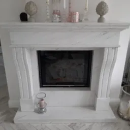 Biały marmurowy kominek z czarnym wkładem kominkowym, ozdobiony dekoracjami w pastelowych kolorach. Widoczne elementy wystroju wnętrza: podłoga w jodełkę, dekoracje na kominku i obok niego.