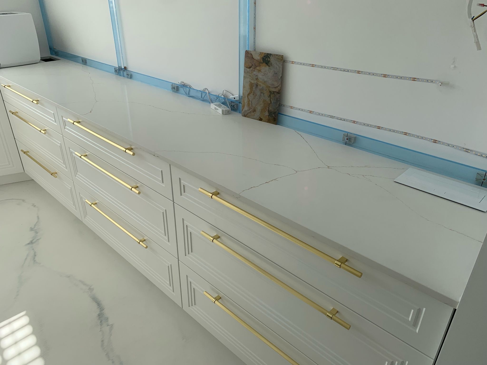 Jasny blat z widocznymi żyłami na białych szafkach z długimi, złotymi uchwytami. Na blacie leży próbka kamienia. W tle widoczne listwy LED i przewody.