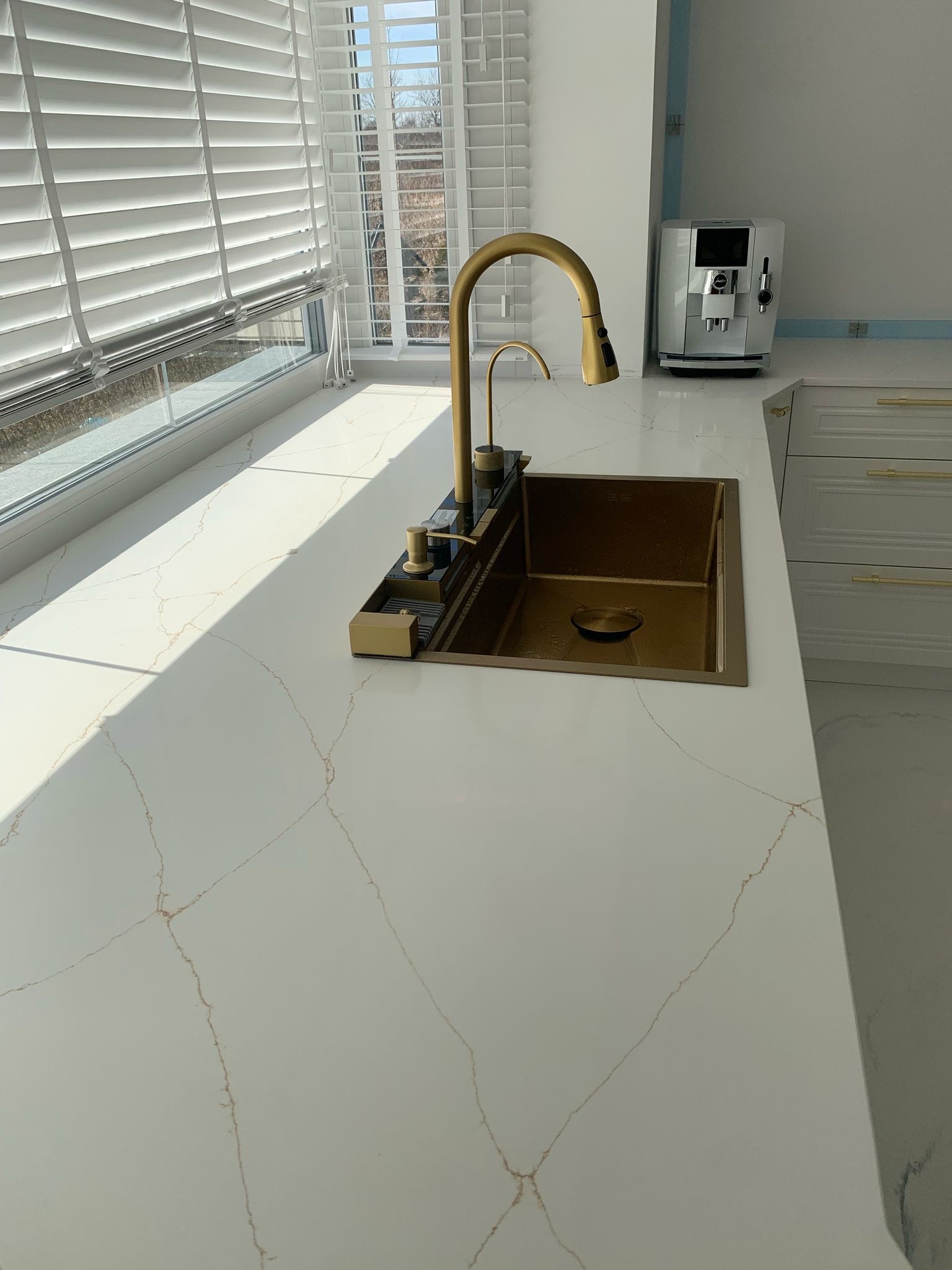 Biały blat kuchenny z subtelnym wzorem żyłkowania w odcieniach złota, złota bateria kuchenna i zlew, białe żaluzje w oknie, ekspres do kawy w tle.