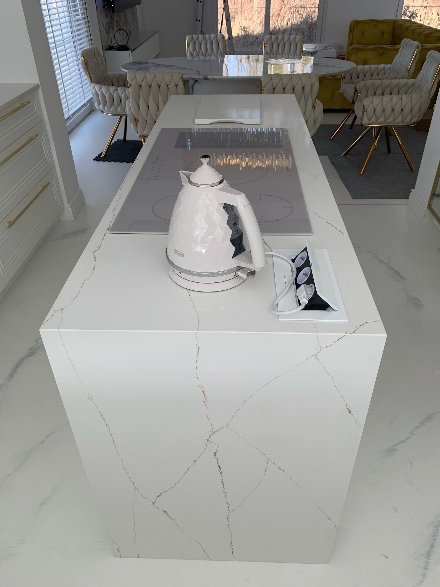 Biały blat kuchenny z konglomeratu kwarcowego z delikatnym żyłkowaniem w odcieniach złota, na którym stoi czajnik elektryczny w stylu retro i wbudowane gniazdka elektryczne.