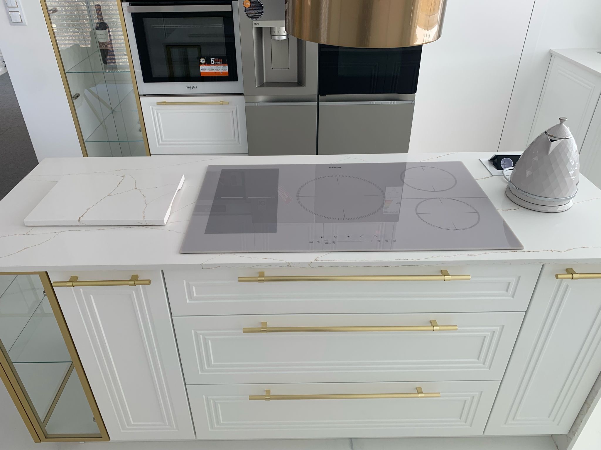 Biały blat kamienny z szarymi żyłami w kuchni, z płytą indukcyjną, złotymi uchwytami szafek i czajnikiem w tle zabudowa kuchenna z piekarnikiem i witryną.