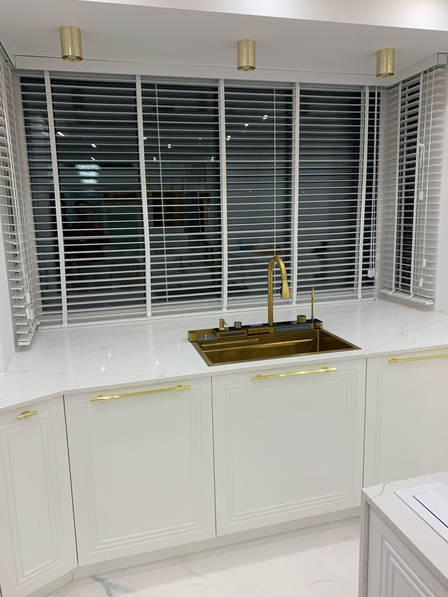 Biała kuchnia z blatem imitującym marmur, zlewem i baterią w kolorze złotym, pod oknem z białymi żaluzjami.