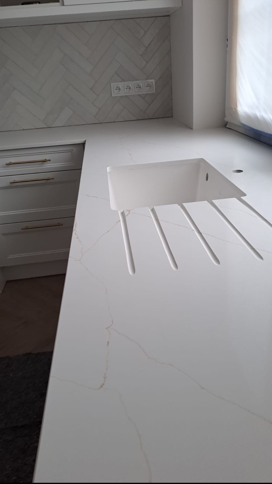 Biały blat z konglomeratu z wbudowanym zlewem o nietypowym kształcie, widoczne złote żyłki na powierzchni, w tle białe szafki i ściana z płytkami ułożonymi w jodełkę.