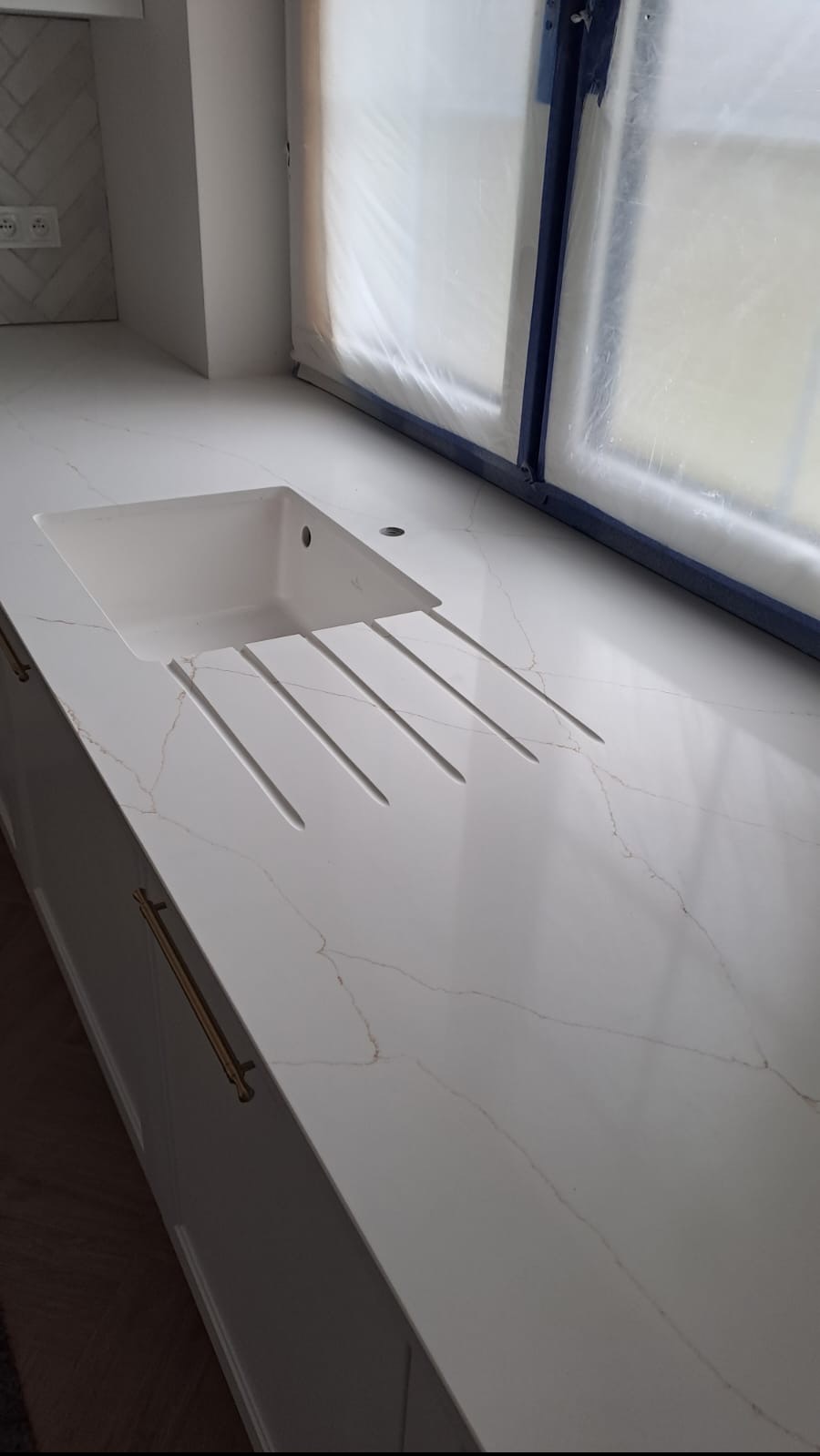 Biały blat kuchenny z konglomeratu z wbudowanym zlewem i ociekaczem, widoczne złote żyłki w strukturze kamienia, okno zabezpieczone folią malarską.