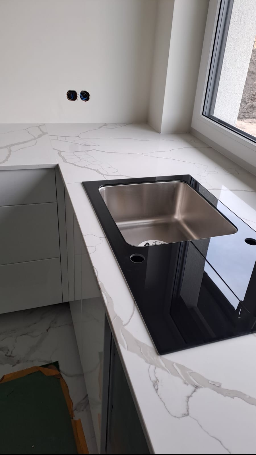 Biały blat kuchenny z szarym żyłkowaniem, z wbudowanym czarnym zlewem z nierdzewnej stali, widoczne gniazdka elektryczne w ścianie, częściowo zdjęta folia ochronna na podłodze.