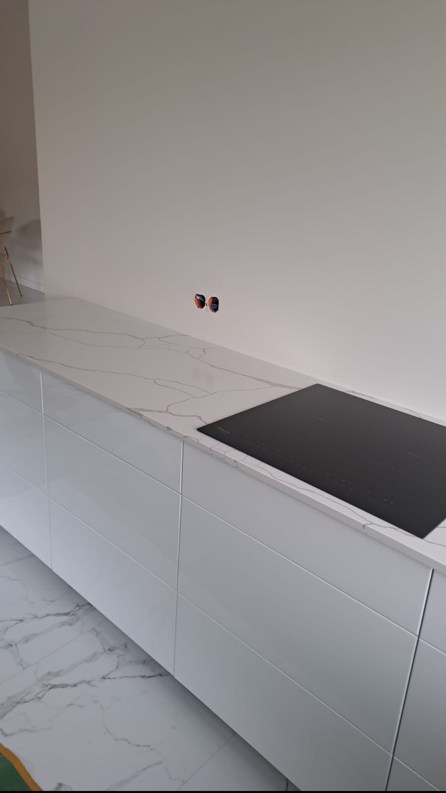 Biały blat kuchenny z marmurkowym wzorem, z wbudowaną czarną płytą indukcyjną, na tle białej ściany z widocznymi gniazdkami elektrycznymi i marmurową podłogą.