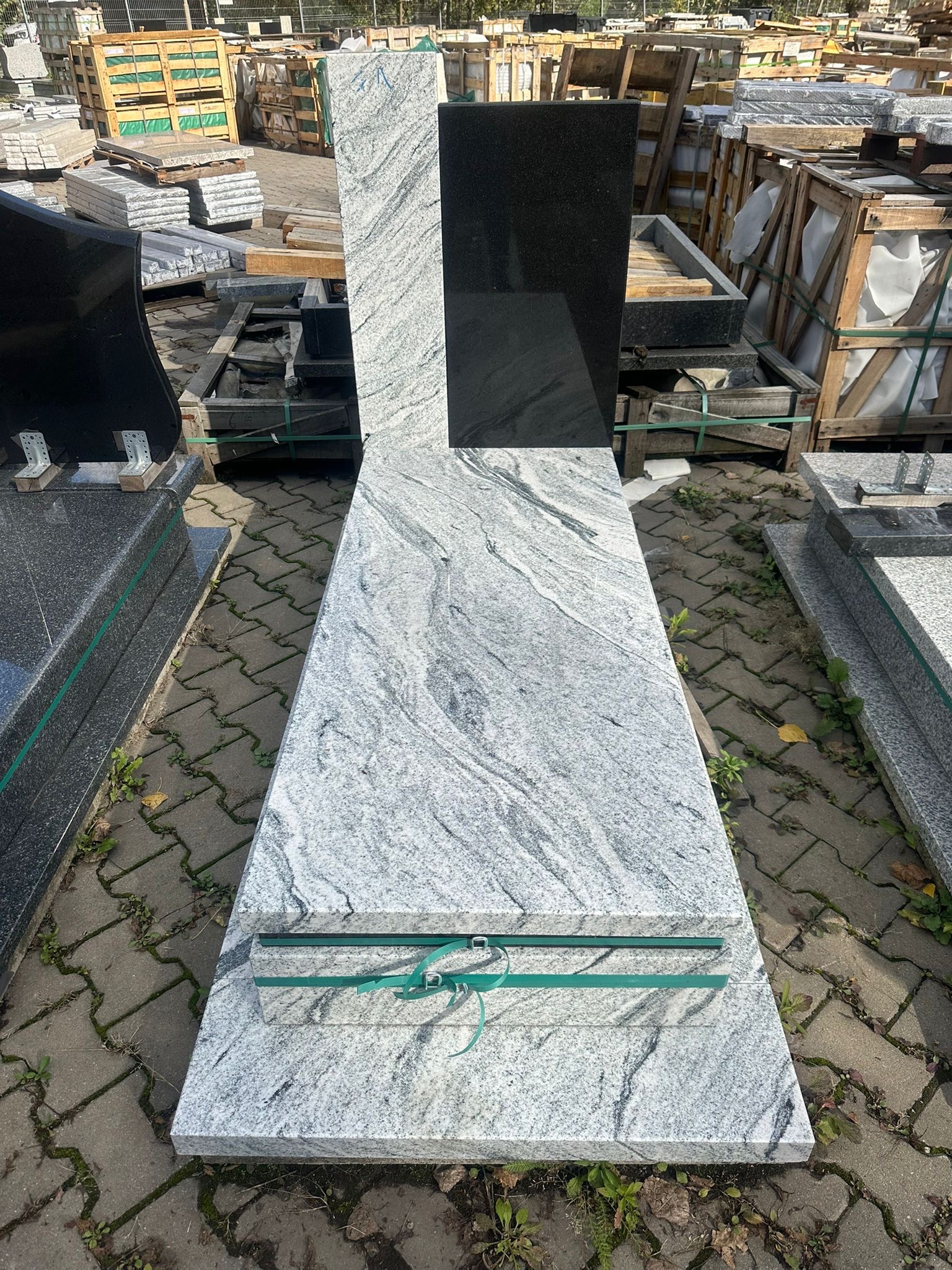 Elementy nagrobka z jasnego, wzorzystego granitu i czarnego kamienia, przygotowane do montażu, zabezpieczone zieloną taśmą, na tle kostki brukowej i palet z innymi elementami kamiennymi.