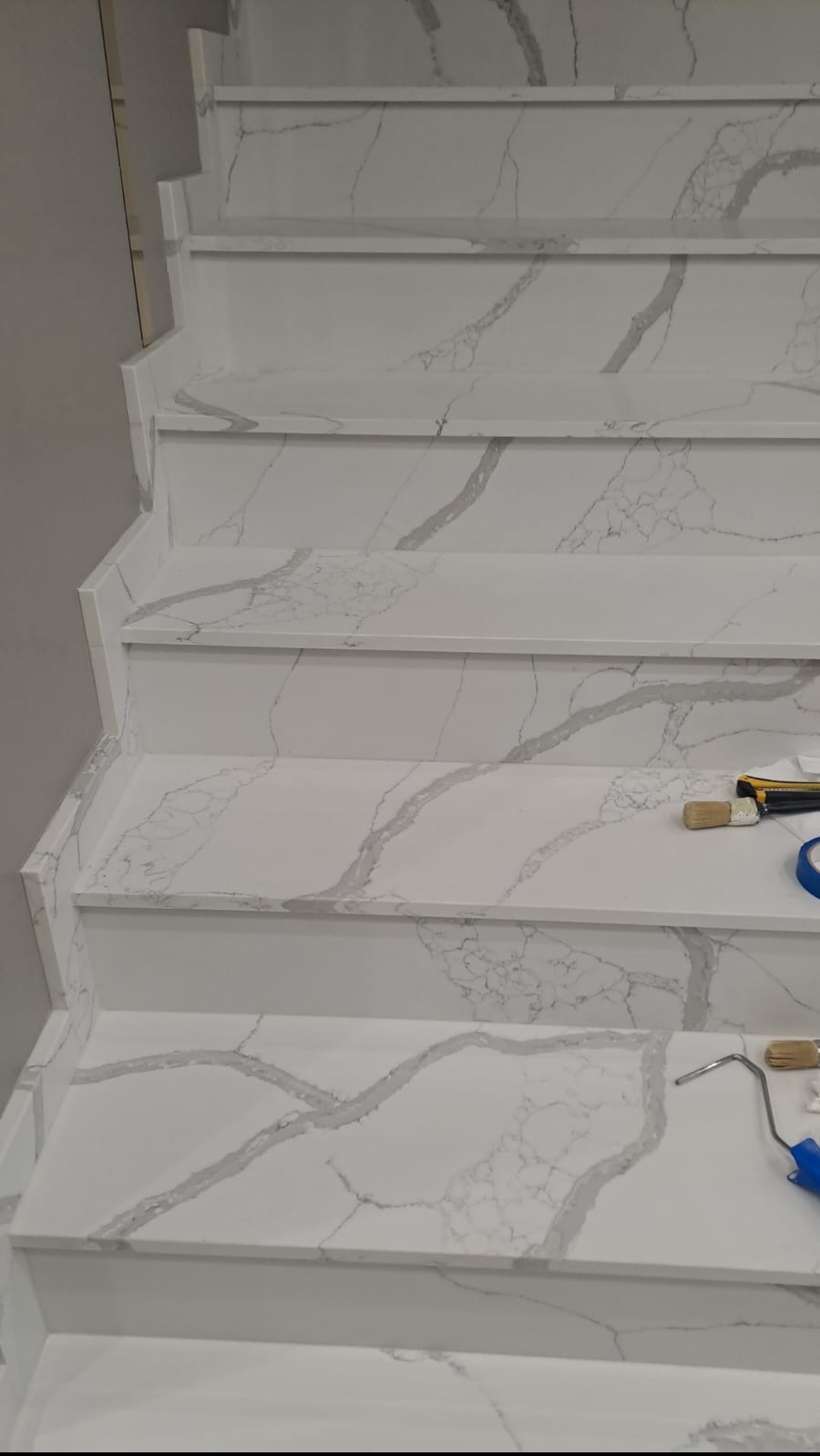 Wykończone schody wewnętrzne z białego kamienia z szarymi żyłami, widoczne narzędzia malarskie i taśma malarska na stopniach.