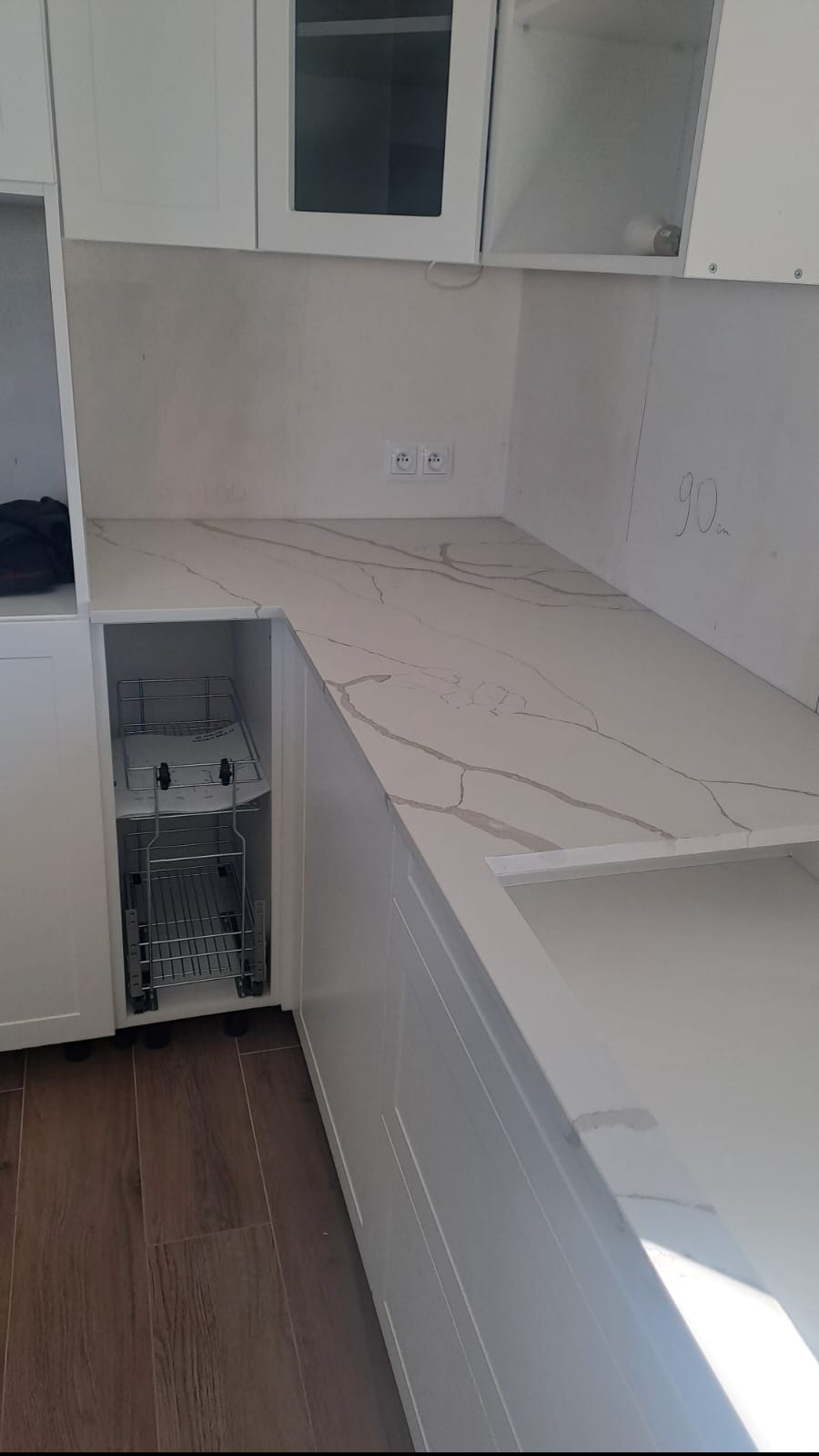 Biały blat kuchenny z marmurkowym wzorem, wbudowany w białe szafki kuchenne z wysuwanym koszem cargo, widoczne gniazdka elektryczne na ścianie.