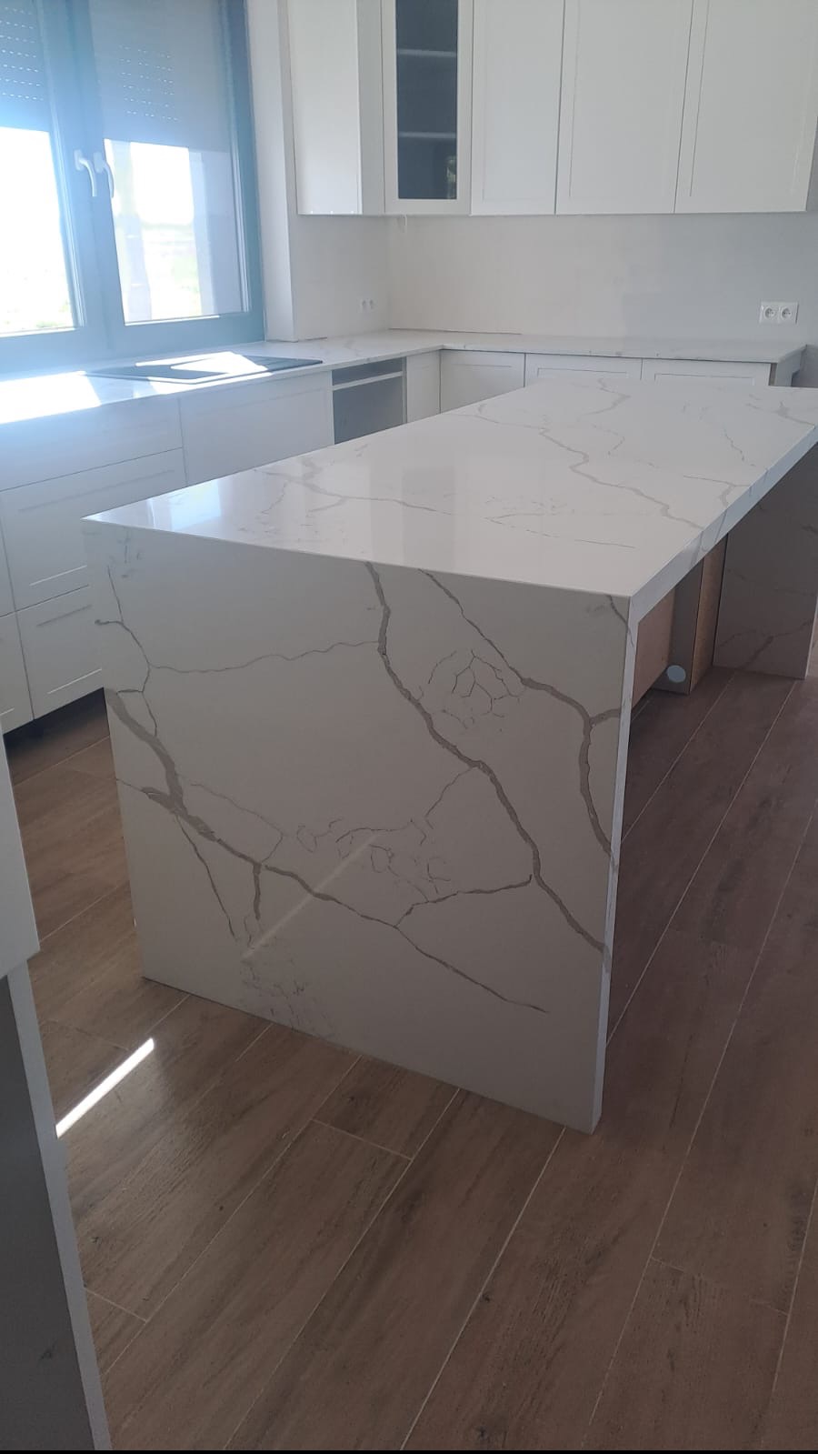Biały, nowoczesny blat kuchenny z charakterystycznym szarym wzorem żyłkowania, stanowiący część wyspy kuchennej w jasnej kuchni z drewnianą podłogą.
