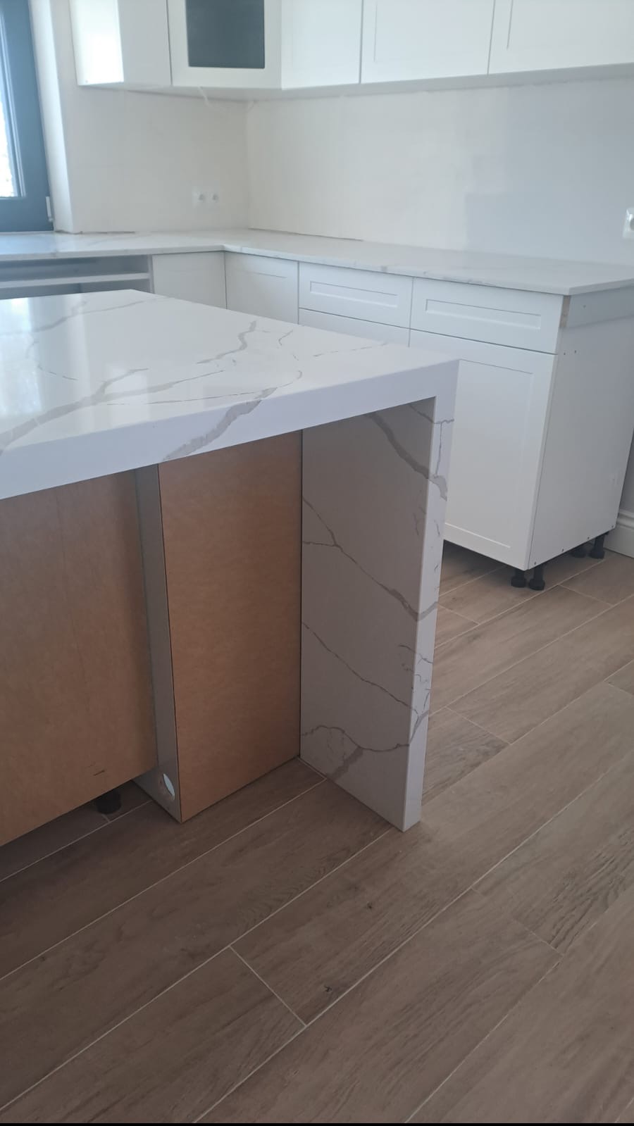 Kuchnia w trakcie montażu: wyspa z białym blatem z wzorem marmuru, widoczne niedokończone elementy zabudowy i drewniane panele boczne. Na podłodze płytki imitujące drewno.