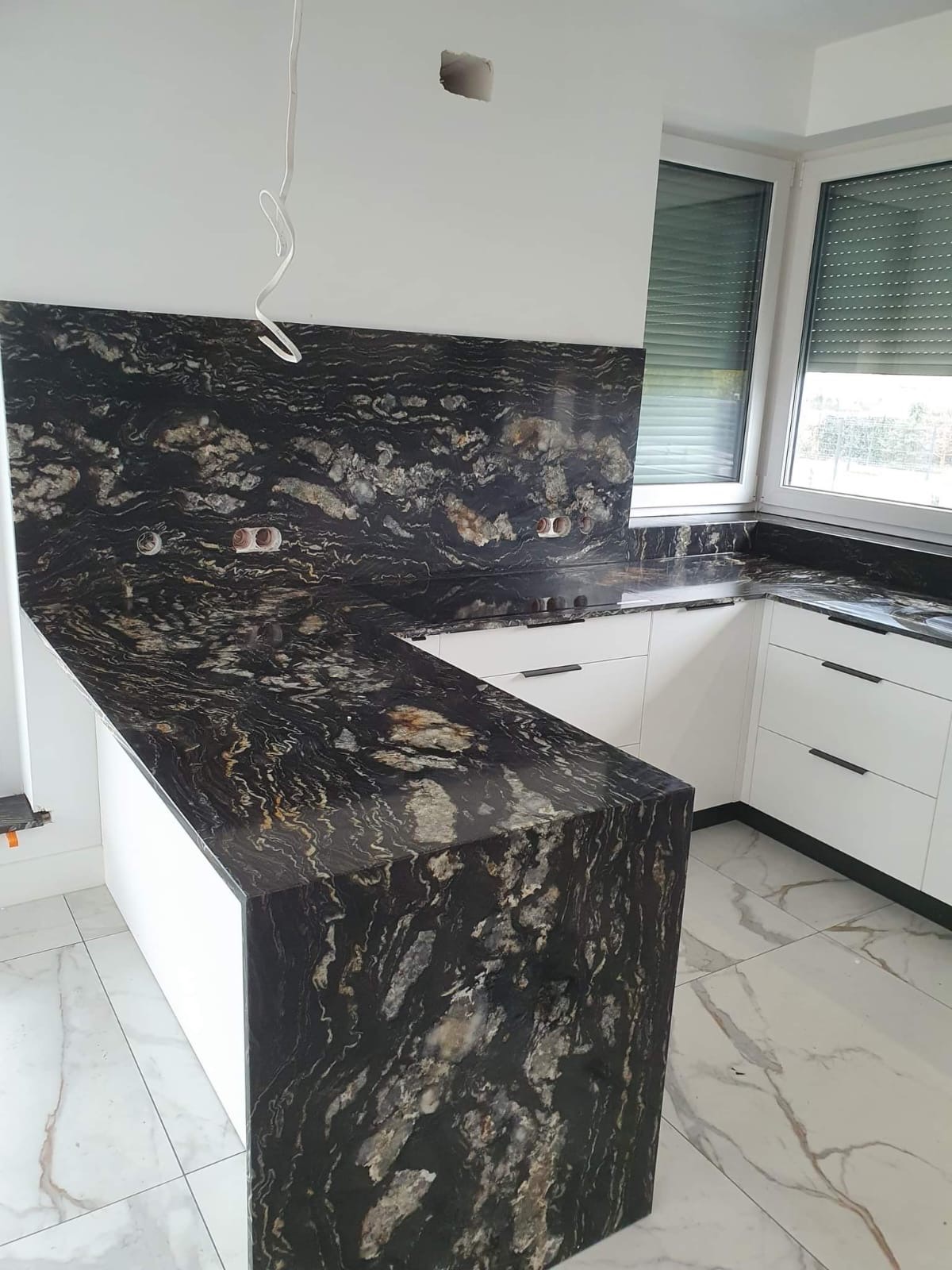 Czarno-złoty blat z kamienia naturalnego, z widocznymi gniazdkami elektrycznymi w ścianie, zamontowany w nowoczesnej kuchni z białymi szafkami i marmurową podłogą.
