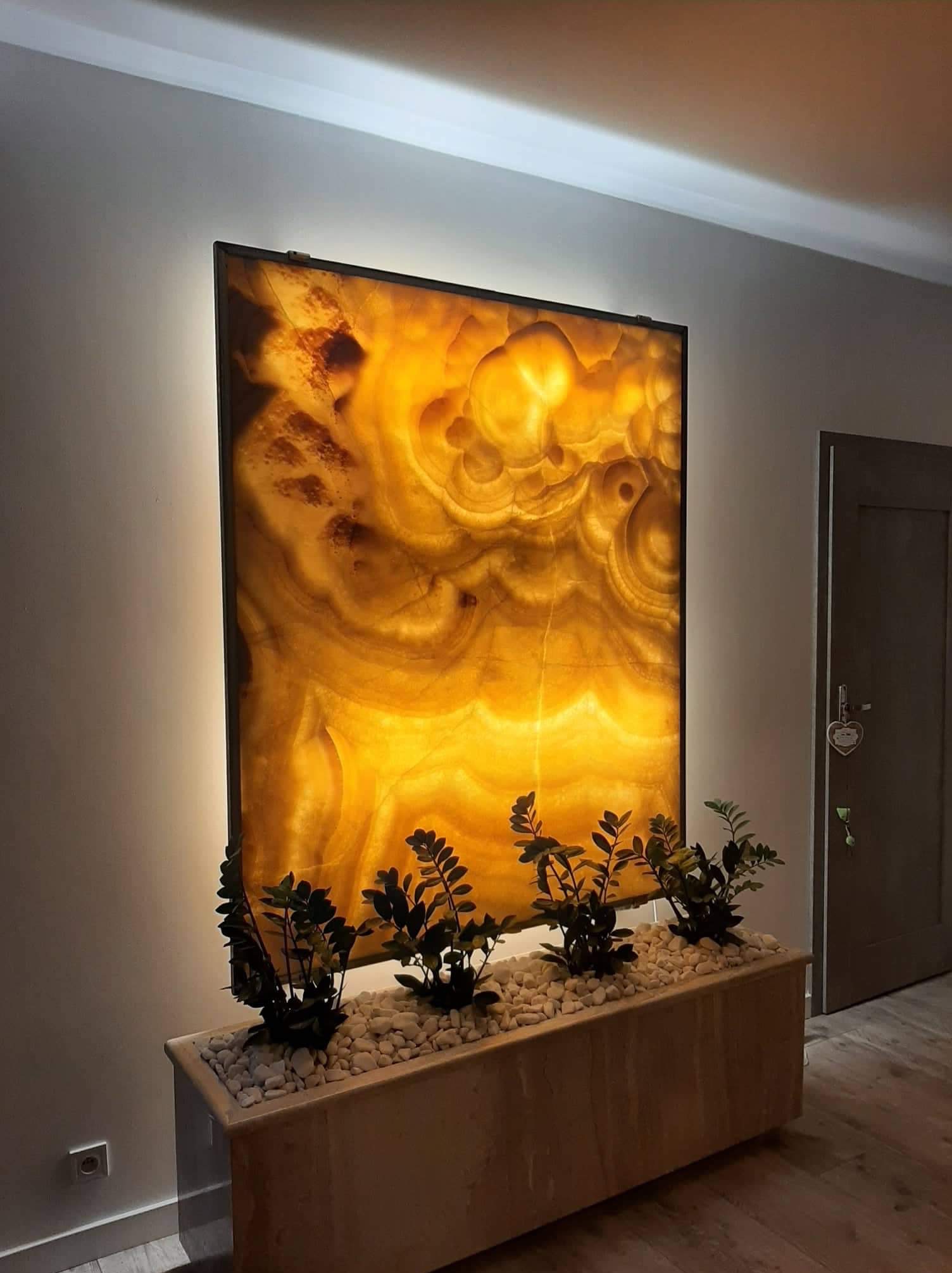 Podświetlany panel z onyksu w czarnej ramie, postawiony na kamiennej donicy z białymi kamykami i zielonymi roślinami, widok na fragment ściany i drzwi.