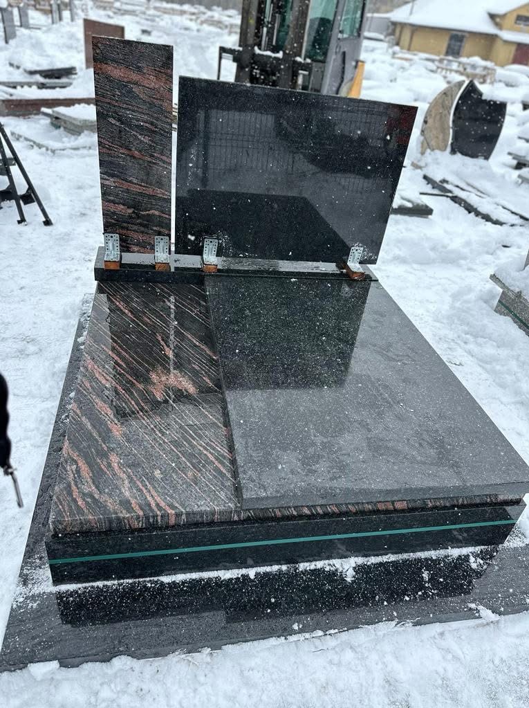 Zimowy kadr nagrobka z ciemnego kamienia z elementami w odcieniach czerwieni, z widocznymi mocowaniami, na tle zaśnieżonego terenu i sprzętu budowlanego.