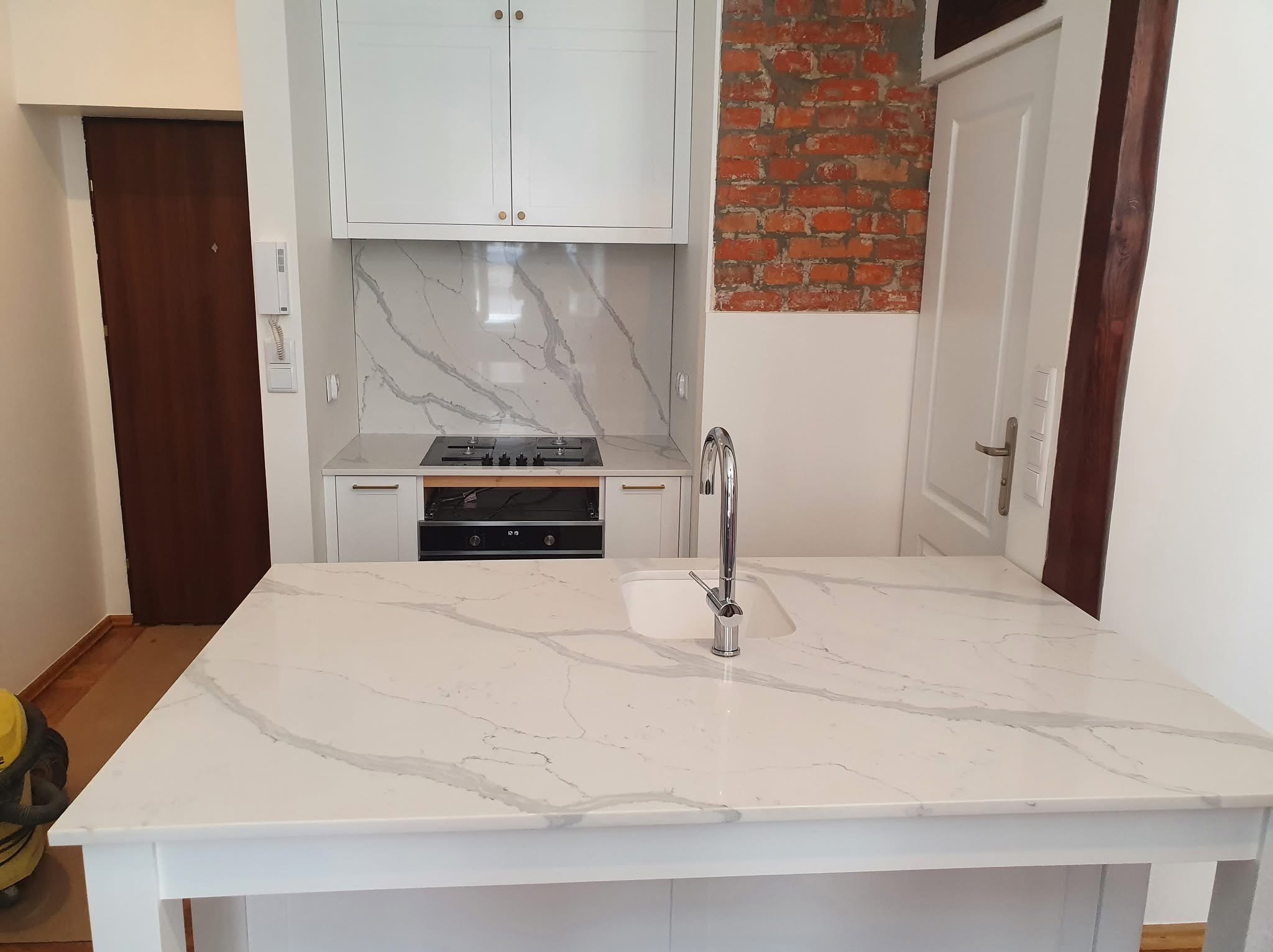 Biały blat kuchenny z marmurowym wzorem na wyspie kuchennej, z widoczną umywalką i baterią. W tle zabudowa kuchenna i fragment ściany z cegły.