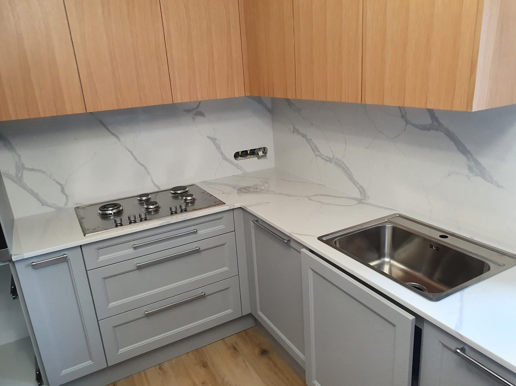 Jasnoszara kuchnia z blatem imitującym marmur, z płytą grzewczą i zlewem. Widoczne drewniane szafki wiszące i szuflady z metalowymi uchwytami.