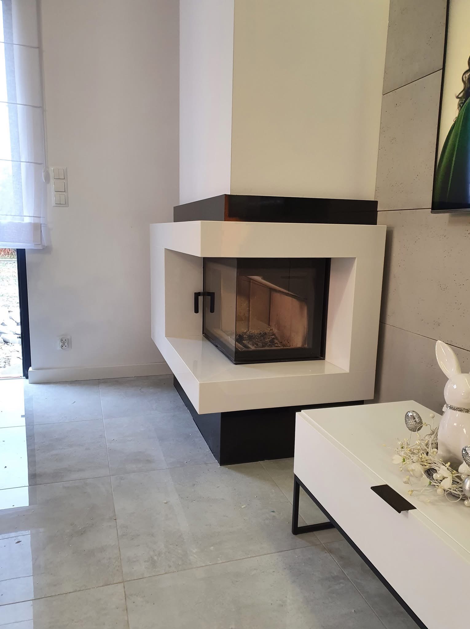 Nowoczesny kominek w salonie z białymi ścianami i szarymi płytkami podłogowymi. Widoczny fragment szafki RTV z dekoracjami wielkanocnymi.