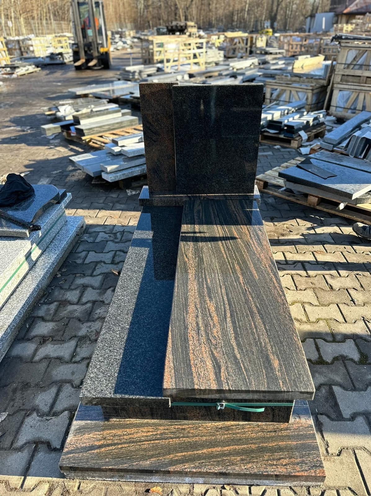 Elegancki nagrobek z ciemnego granitu z wyraźnym wzorem słojów, ustawiony na zewnątrz, w tle inne elementy kamieniarskie i wózek widłowy.