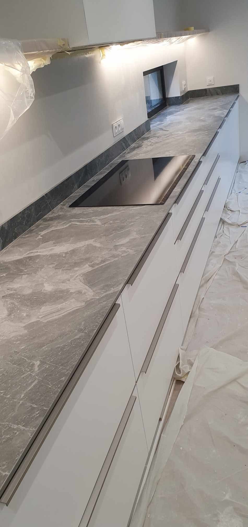 Szary blat kamienny z wbudowaną płytą indukcyjną, białe szafki z długimi uchwytami, osłonięte folią malarską. Widok na fragment kuchni po montażu.