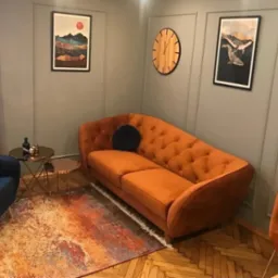 Pomarańczowa sofa z weluru z pikowaniem w gabinecie z szarymi ścianami, drewnianym zegarem i obrazami w czarnych ramach.